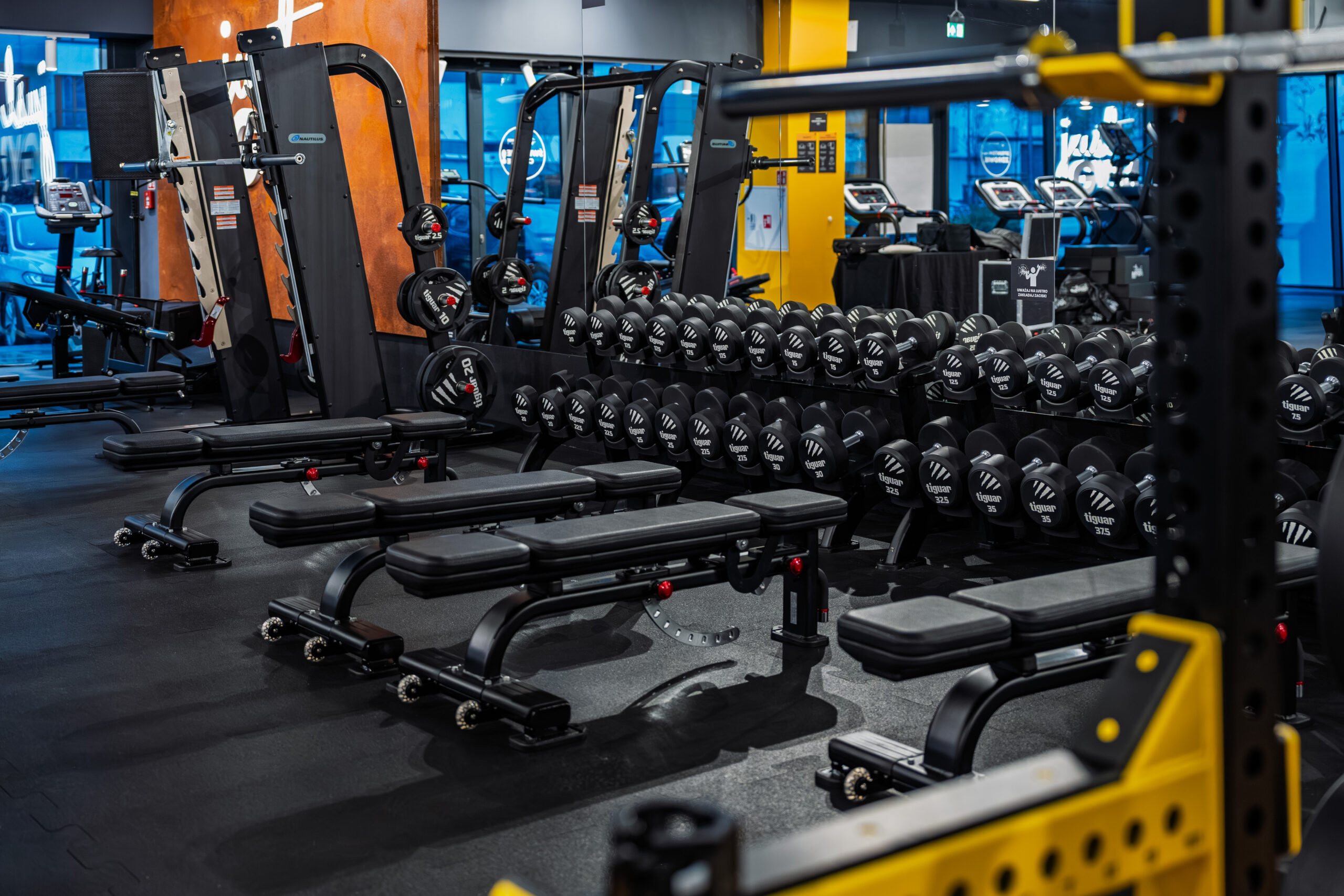 Strefa wolnych ciężarów w siłowni na ursusie Just GYM BOX z hantlami na stojakach, ławkami treningowymi i maszynami siłowymi.