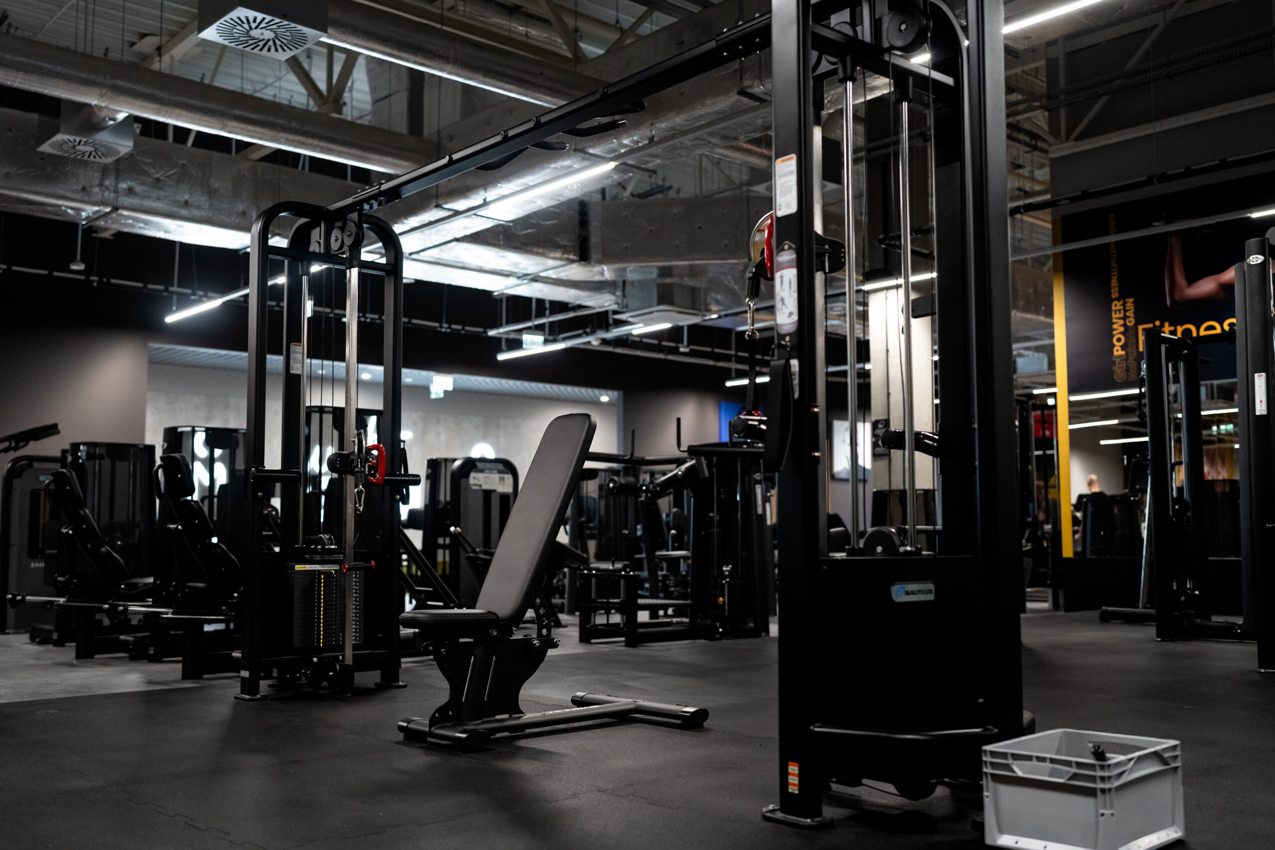 Wnętrze nowoczesnej siłowni z maszynami do ćwiczeń siłowych z obciążeniem stosowym i suwnicą bramową. Widoczna jest otwarta przestrzeń z czarną gumową podłogą, betonowym sufitem i odsłoniętymi instalacjami wentylacyjnymi.