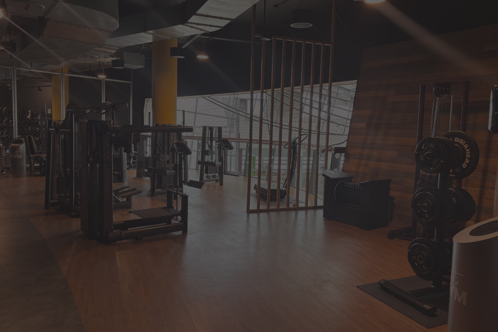Зона силових тренувань у клубі Just GYM Гдиня Батори, з помітним стійкою для олімпійських ваг, тренажерами, дерев’яними стіновими панелями та декоративною ажурною перегородкою, виконана в сучасному індустріальному стилі.