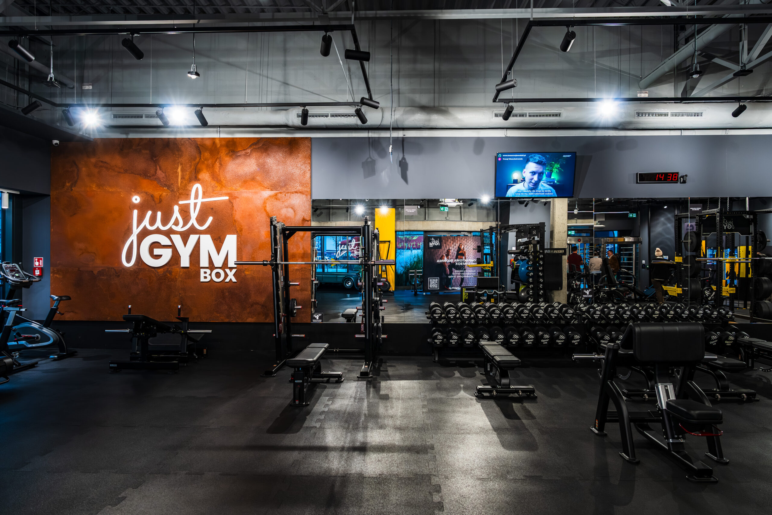 Інтер'єр тренажерного залу в районі Урсус у Варшаві Just GYM BOX з логотипом клубу, зоною вільних ваг та сучасним освітленням.