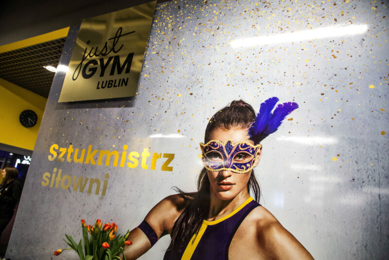 The entrance to the Just Gym Lublin with promotional graphics and the words „Gym master” against a złoto-grey wall.