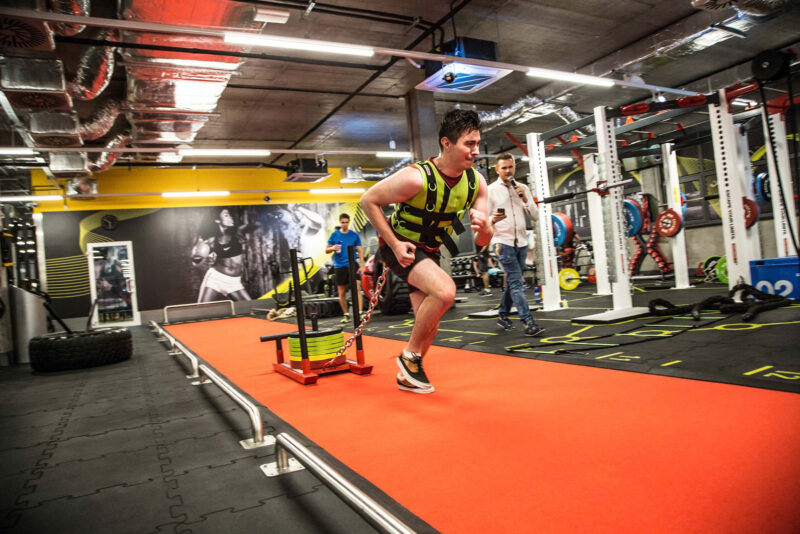 Mężczyzna wykonujący sprint z obciążeniem na czerwonym torze treningowym w siłowni Just Gym Kraków – strefa treningu funkcjonalnego.