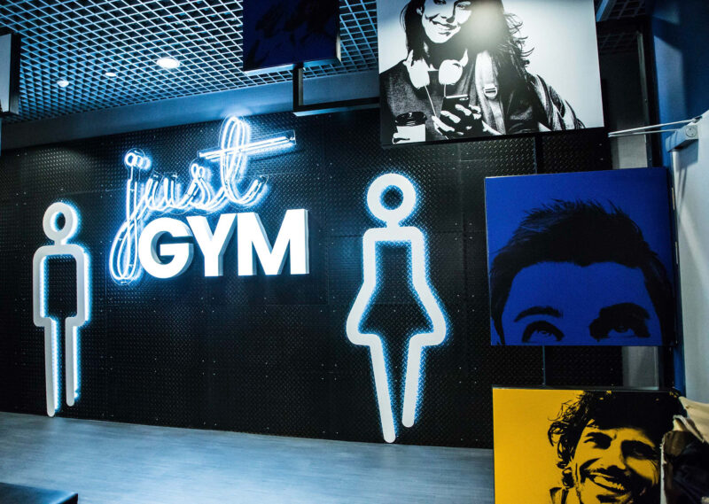 Wejście do siłowni Just Gym Kraków – neonowe logo klubu z ikonami postaci i grafikami na ścianie.