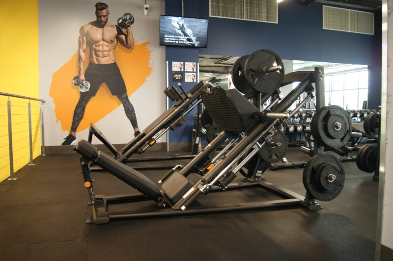 Gym Ursynów Warsaw and leg machines in the gym's strength area with a large mural depicting an athlete holding dumbbells.