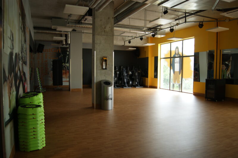 Gym Ursynów Warsaw - Fitness room with mirrors, yellow walls and wooden floor, equipped with step platforms and group exercise equipment.