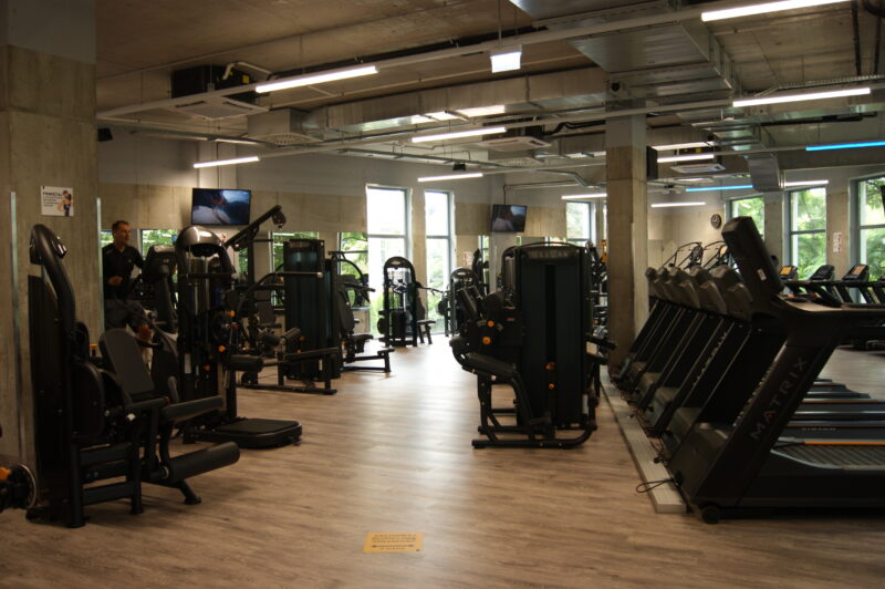 Modern Ursynów Warsaw gym with weight training machines, barbell racks and colourful fitness balls by large windows.