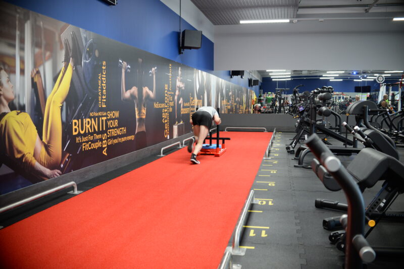 Trening siłowy w siłowni Just Gym Jastrzębie-Zdrój – mężczyzna wykonujący ćwiczenie z wózkiem treningowym na czerwonym torze funkcjonalnym.