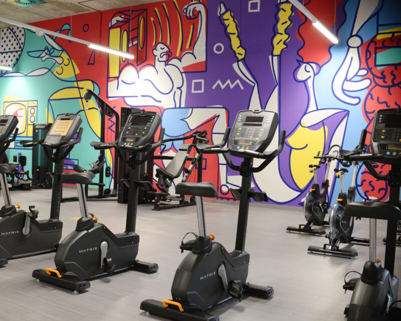 A colourful fitness zone at Just Gym Łódź Rembielińskiego with stationary bikes and an artistic mural on the wall.