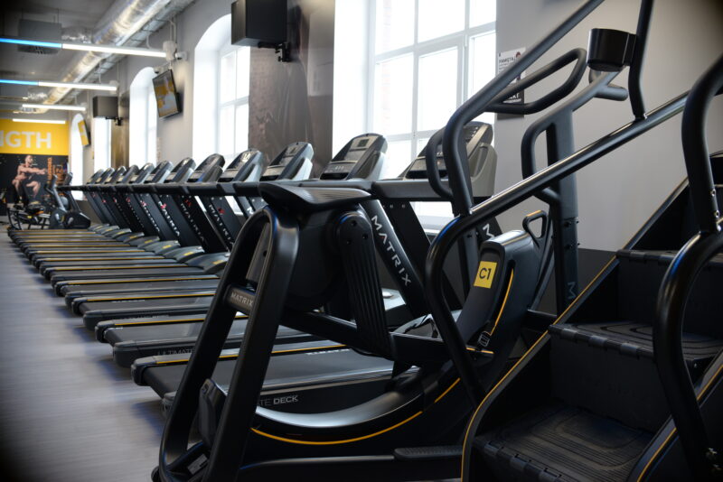 Strefa cardio w Just Gym Racibórz – rząd bieżni i schodów treningowych Matrix przy dużych oknach klubu.