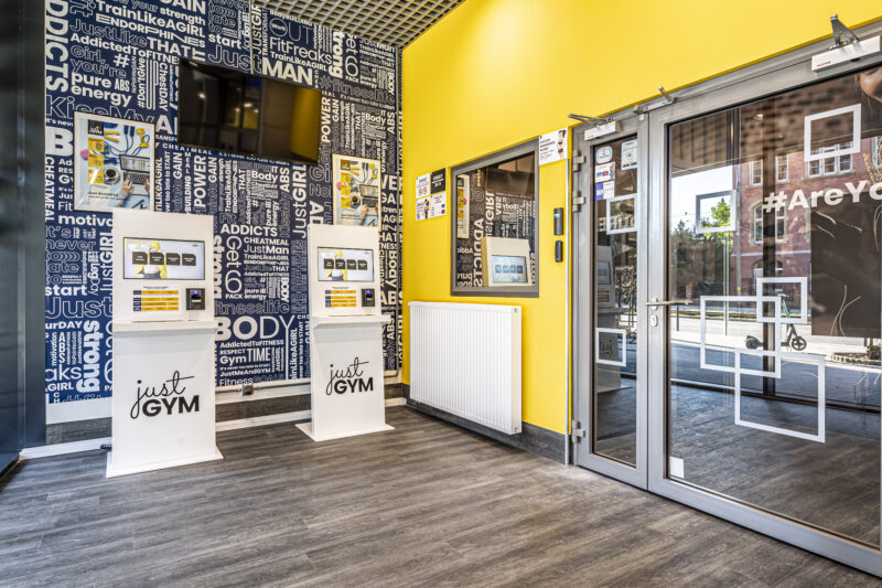 Вхід до Just Gym Wrocław з кіосками самообслуговування та жовтою і темно-синьою стіною, прикрашеною мотиваційними фітнес-слоганами.
