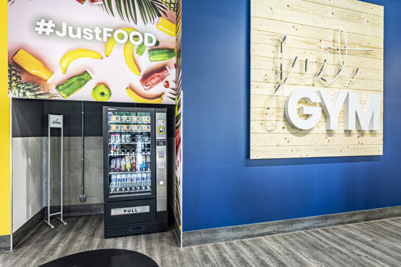 Автомат з продажу напоїв і закусок JustFood у спортзалі Just Gym Wrocław з барвистим зображенням фруктів і логотипом спортзалу на синій стіні.
