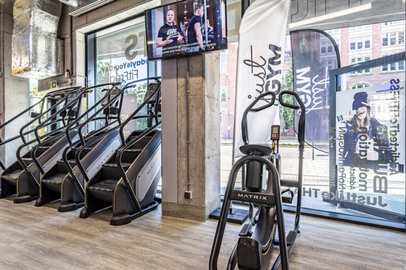 Кардіо-зона Just Gym Wrocław з біговими доріжками та тренувальними сходами Matrix біля великих вікон.