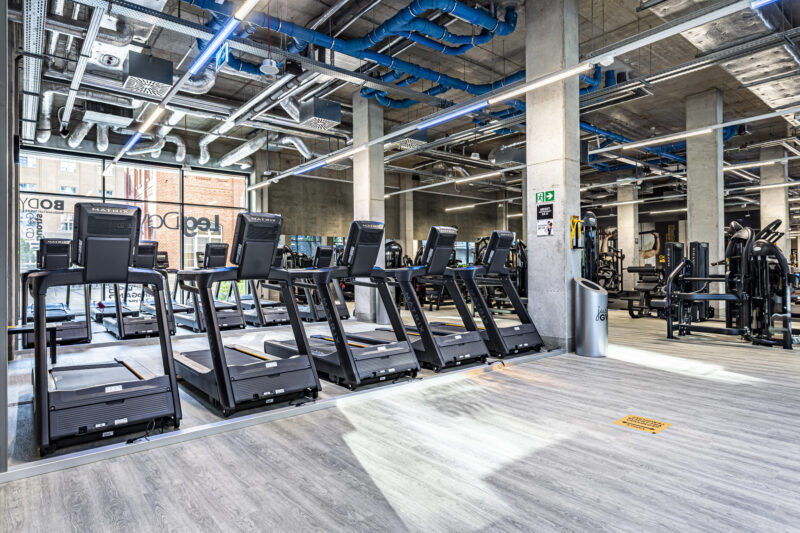 Ряд бігових доріжок у Just Gym Wrocław з видом на місто крізь скляні стіни, в індустріальному інтер'єрі з бетонними колонами.