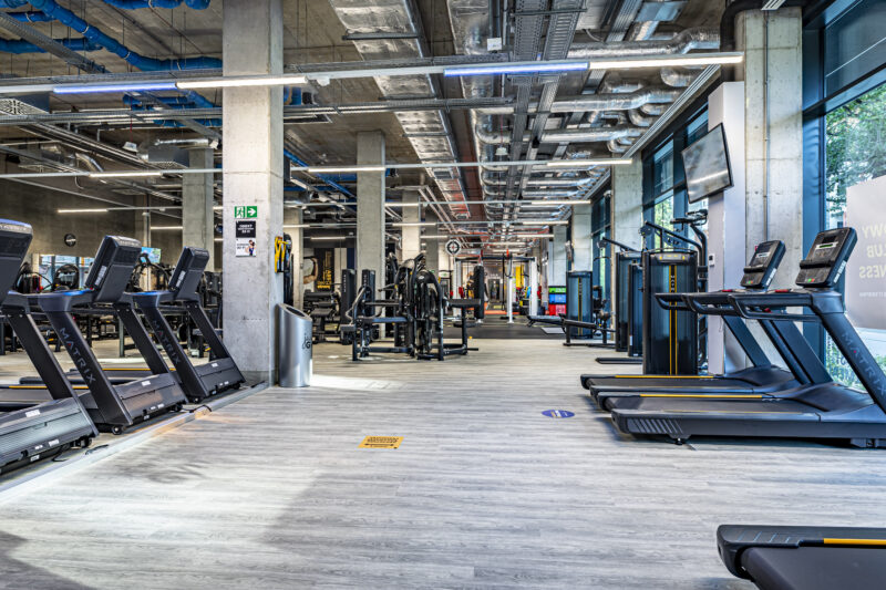 Панорамний вид на тренувальну зону Just Gym Wrocław з біговими доріжками, силовими тренажерами та яскравим індустріальним дизайном.