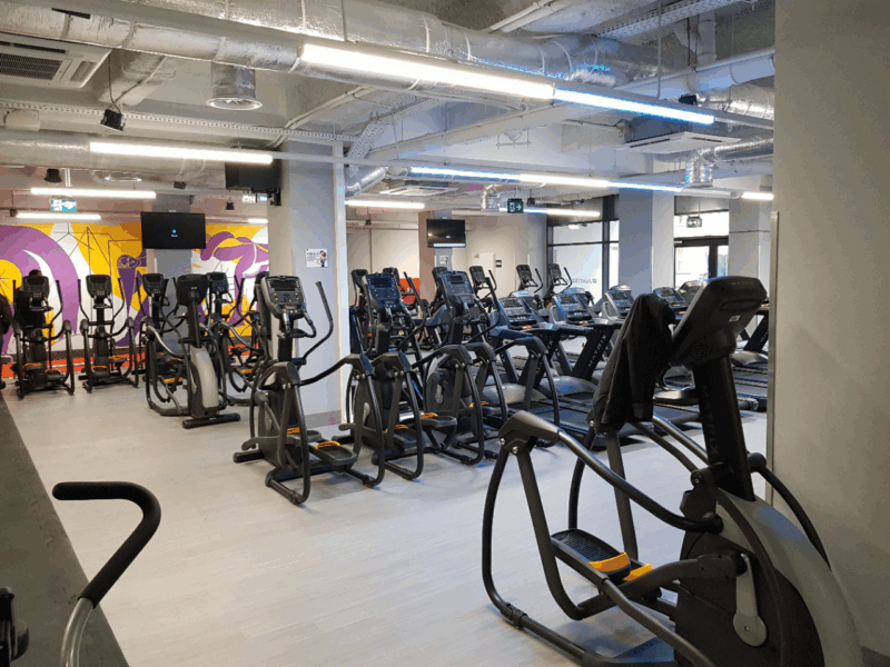 Strefa cardio w siłowni Just Gym Łódź Rewolucji z bieżniami i orbitrekami na tle kolorowego muralu fitness.