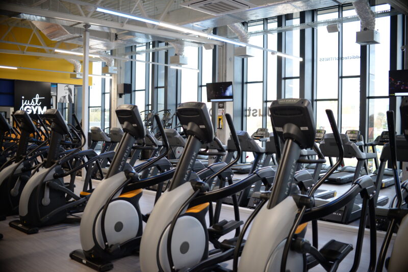 Nowoczesna siłownia z równymi rzędami maszyn Matrix – orbitreków, rowerów i bieżni, w tle logo Just Gym na żółtej ścianie.