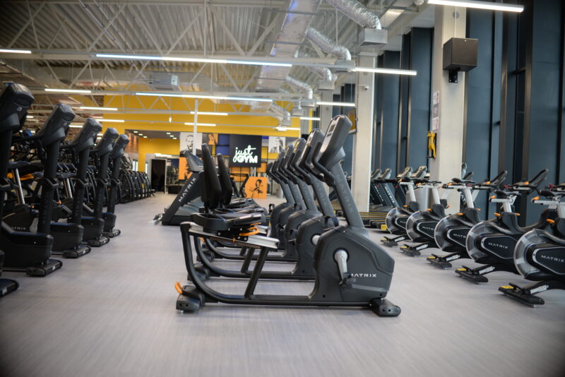 Rząd orbitreków i bieżni w nowoczesnej siłowni Just Gym z dużymi przeszkleniami i żółtymi akcentami na ścianach.