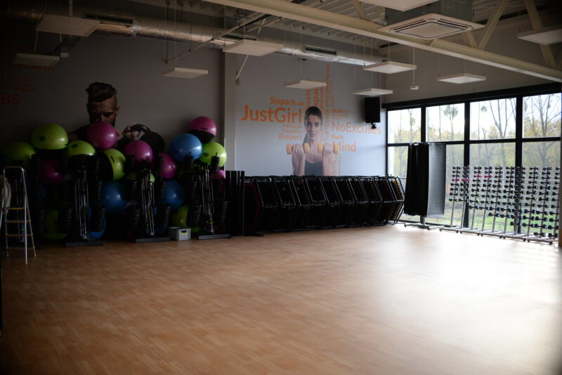 Jasna sala fitness z dużymi oknami, kolorowymi piłkami gimnastycznymi i sprzętem do zajęć grupowych ustawionym pod ścianą z motywacyjnymi grafikami.