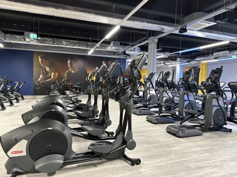A modern cardio zone in the Just Gym Częstochowa gym with rows of orbiters and exercise bikes, against a wall with motivational graphics of athletes.