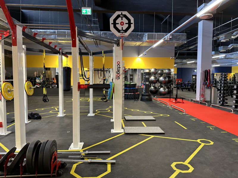 Functional zone in the Just Gym Częstochowa gym with training structure, weights, TRX and boxing bag, with yellow and black signage on the floor.