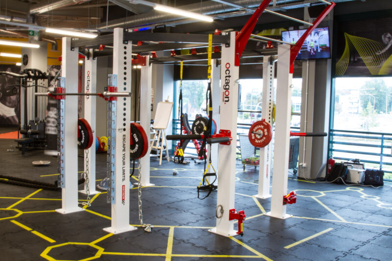 Just Gym Plock functional training zone - barbells, weights and exercise accessories in a space with yellow markings on the floor.
