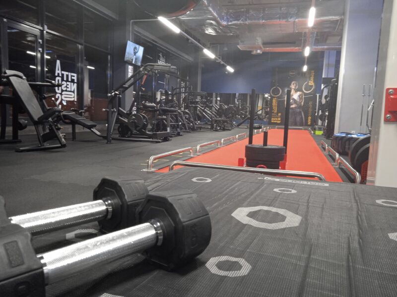 A strength area with benches, dumbbells and exercise machines, dumbbells on a mat in the foreground and a red training track in the background.