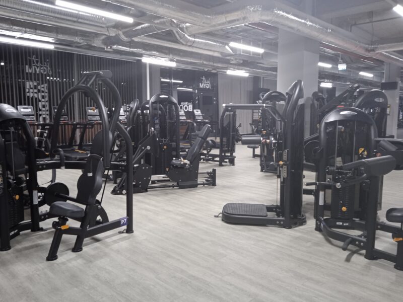 Gym area with numerous weight training machines, well lit, with visible treadmills by the windows.