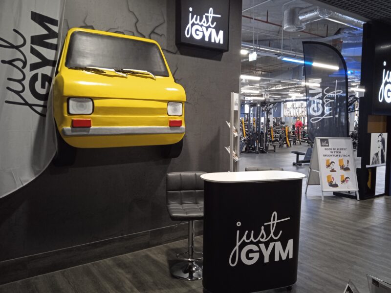 The gym reception area with the „Just Gym” logo, a yellow model car on the wall and a view of the training room in the background.