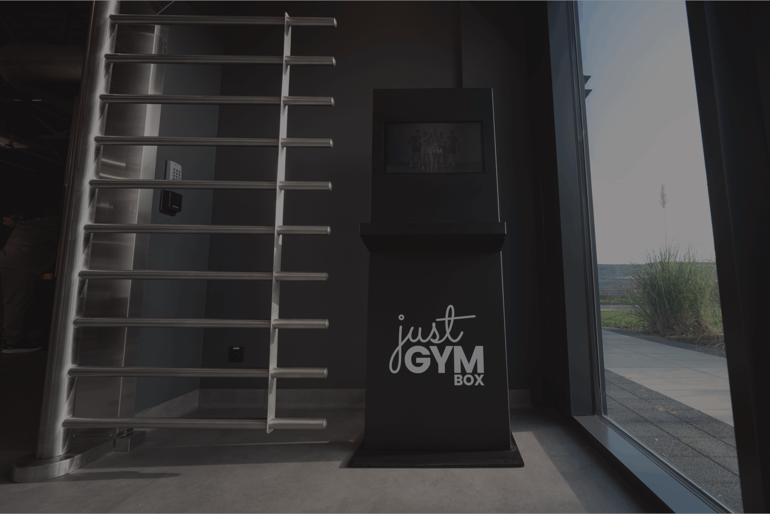 Bramka wejściowa i recepcja samoobsługowa w Just Gym Box Warszawa Herbu Oksza – kołowrotek, terminal z logo klubu przy przeszklonym wejściu.