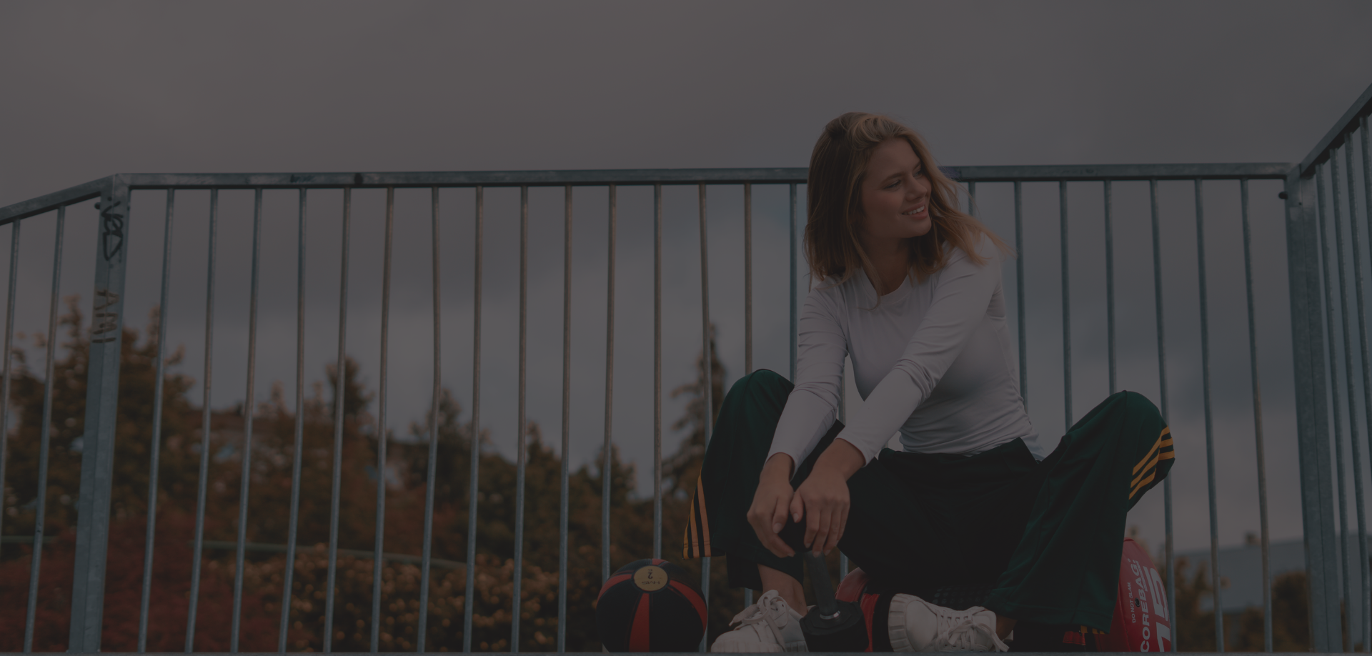 Uśmiechnięta kobieta w stroju sportowym siedzi na zewnętrznej rampie, obok piłki lekarskiej i torby treningowej.