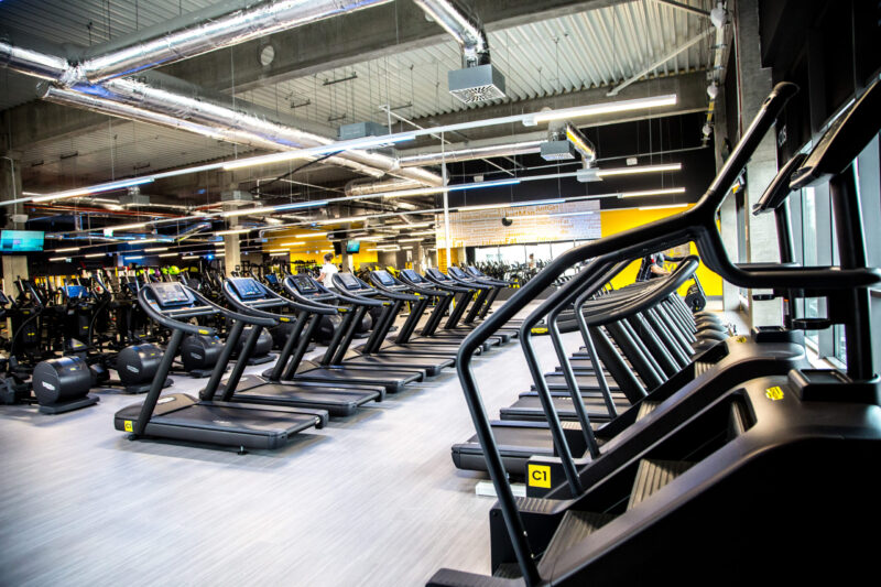 Jasna, przestronna sala cardio z licznymi bieżniami Technogym ułożonymi w rzędach, z widoczną industrialną instalacją pod sufitem