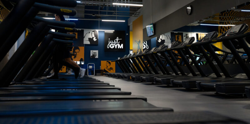 Rząd nowoczesnych bieżni w klubie Just GYM na tle ciemnoniebieskiej ściany z grafikami motywacyjnymi i dużym logo sieci