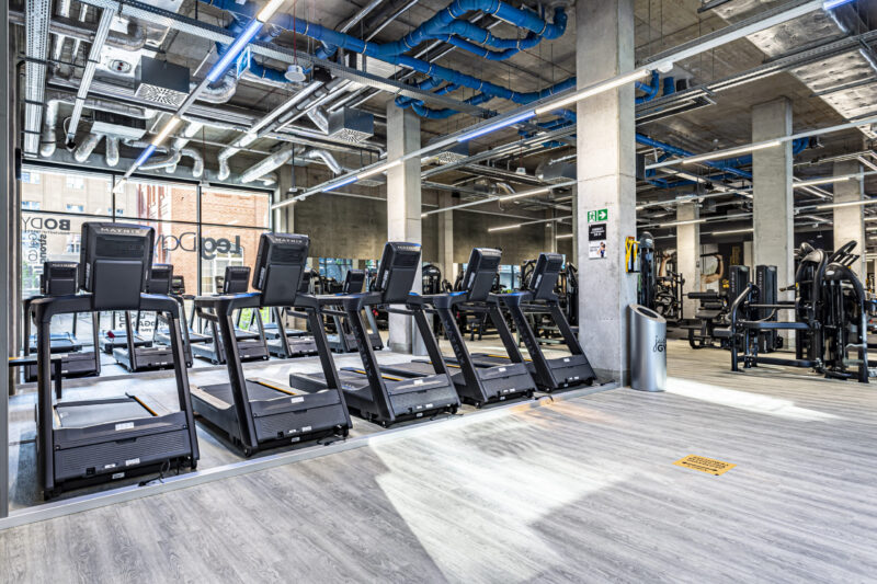 Nowoczesna strefa cardio z bieżniami Matrix w industrialnym wnętrzu z widocznymi niebieskimi rurami wentylacyjnymi i betonowymi filarami