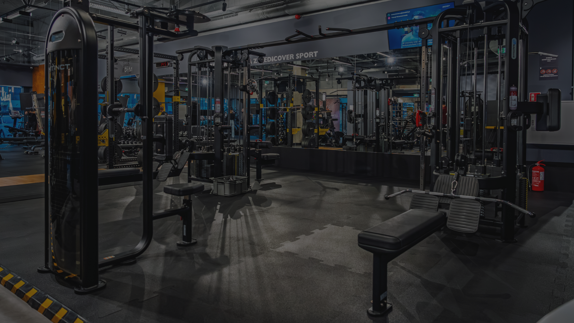 Szerokie ujęcie wnętrza nowoczesnej siłowni Just GYM BOX, pokazujące strefę maszyn siłowych, stojaki z hantlami oraz ławki treningowe. W tle widać ściany z grafikami oraz oświetlenie typu track.