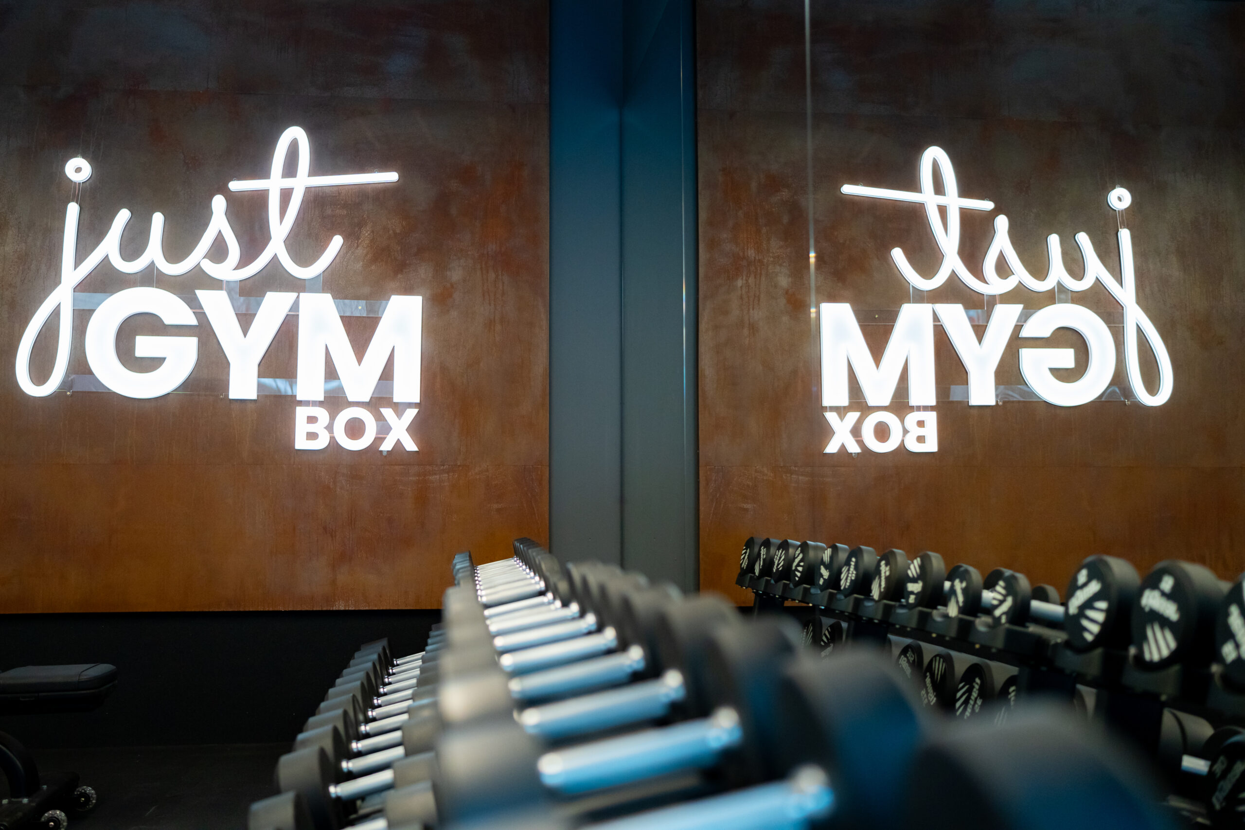 Крупним планом неоновий логотип "just GYM BOX" на стіні з текстурою іржі, відбитий у дзеркалі поруч з рядами гантелей.